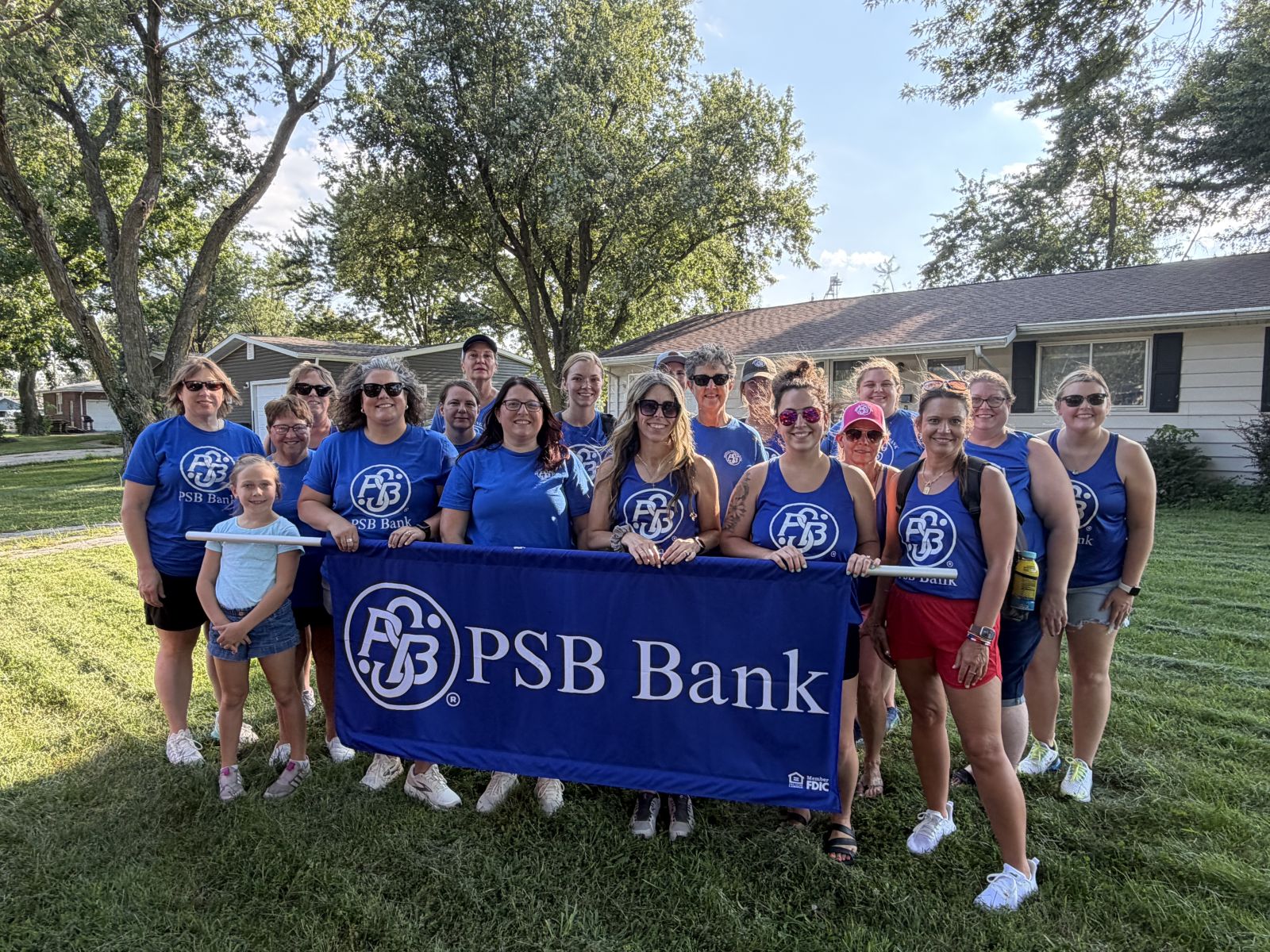 PSB Bank employees and community members pose together outdoors in matching blue PSB Bank shirts while holding a large PSB Bank banner.